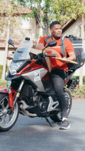 A delivery rider on a motorcycle holding a package outdoors in a suburban setting.